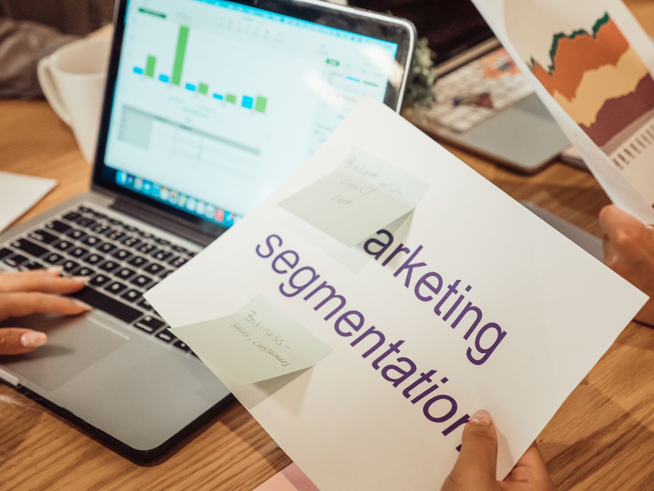 A contemporary workspace featuring hands reviewing marketing data on a laptop and printed documents.